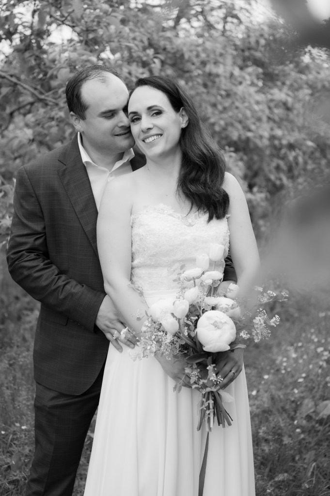 Un couple amoureux en noir et blanc, le marié embrassant la mariée sur l'épaule. La mariée porte une robe blanche et tient un bouquet de fleurs, souriant à l'appareil photo, tandis que le marié la serre doucement.