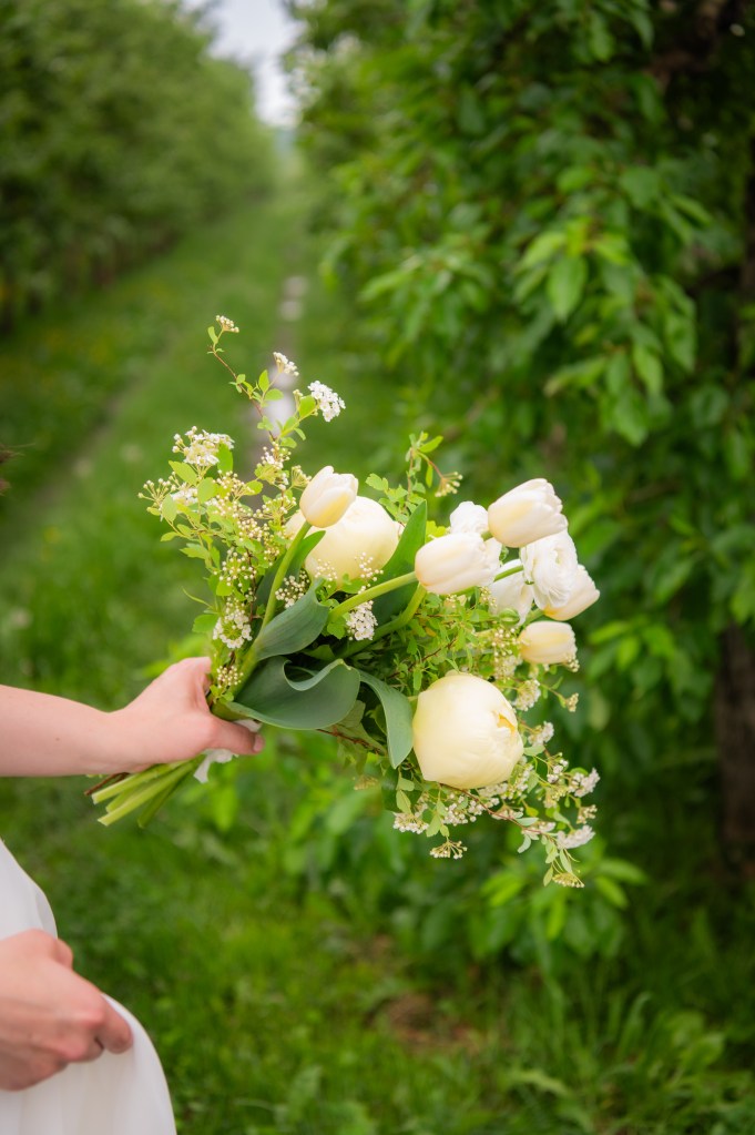 Bouquet de fleurs blanches et vertes tenu par une main, avec un sentier herbeux bordé d'arbres en arrière-plan.