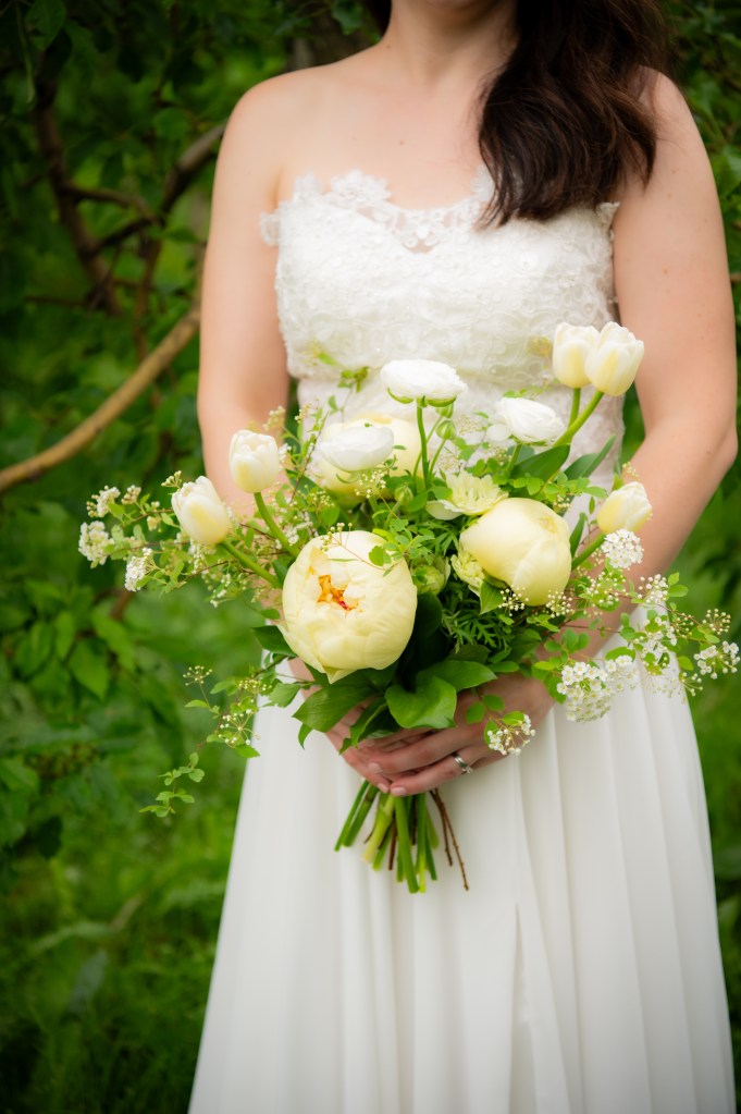 Une femme portant une robe de mariée blanche tient un bouquet de fleurs blanches et jaunes dans un environnement verdoyant.
