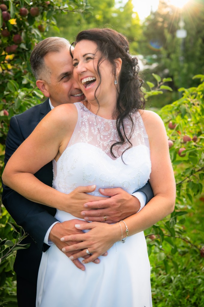 Un couple heureux se tenant dans un verger, le marié embrassant tendrement la mariée qui rit aux éclats, vêtue d'une robe de mariée blanche.