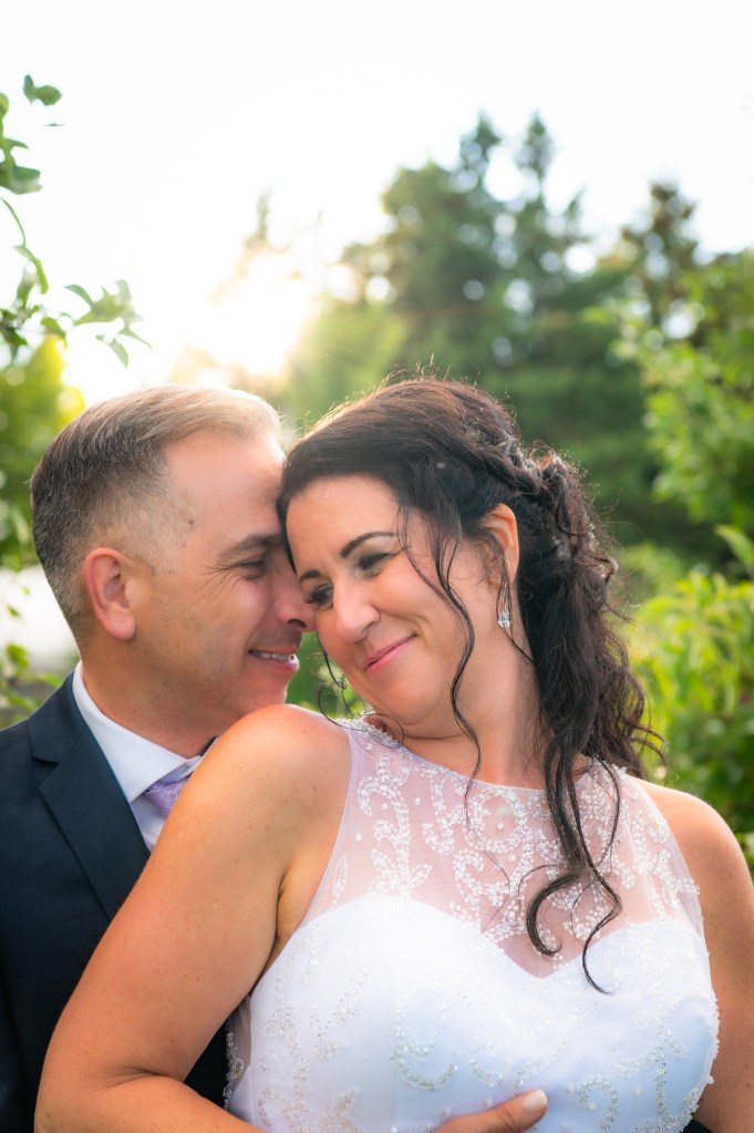 Un couple heureux se regardant tendrement, le marié portant un costume et la mariée une robe de mariée ornée de perles, dans un cadre naturel ensoleillé.