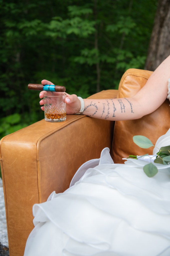Une femme en robe de mariée tient un verre avec une boisson et un cigare sur un canapé en cuir, entourée par la nature.