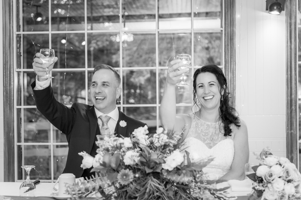 Un couple célèbre en levant leurs verres lors d'un toast, entouré de fleurs sur une table, avec de grandes fenêtres en arrière-plan.