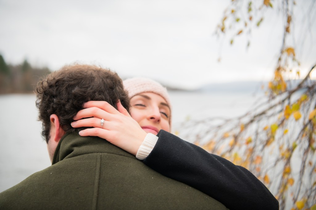 Un couple s'embrasse tendrement près d'un lac, avec des feuilles d'automne et un ciel nuageux.