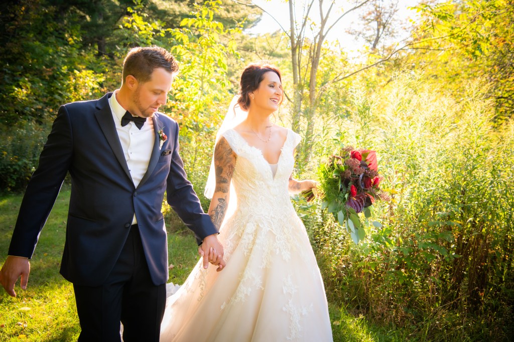 Un couple de jeunes mariés marchant ensemble dans un parc ensoleillé, la mariée portant une robe blanche et tenant un bouquet, tandis que le marié en costume noir les accompagne.