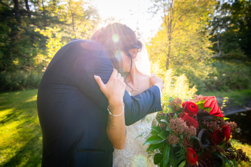 Un couple de fiancés s'enlace dans un jardin ensoleillé, le marié en costume bleu et la mariée en robe blanche tenant un bouquet de fleurs rouges.
