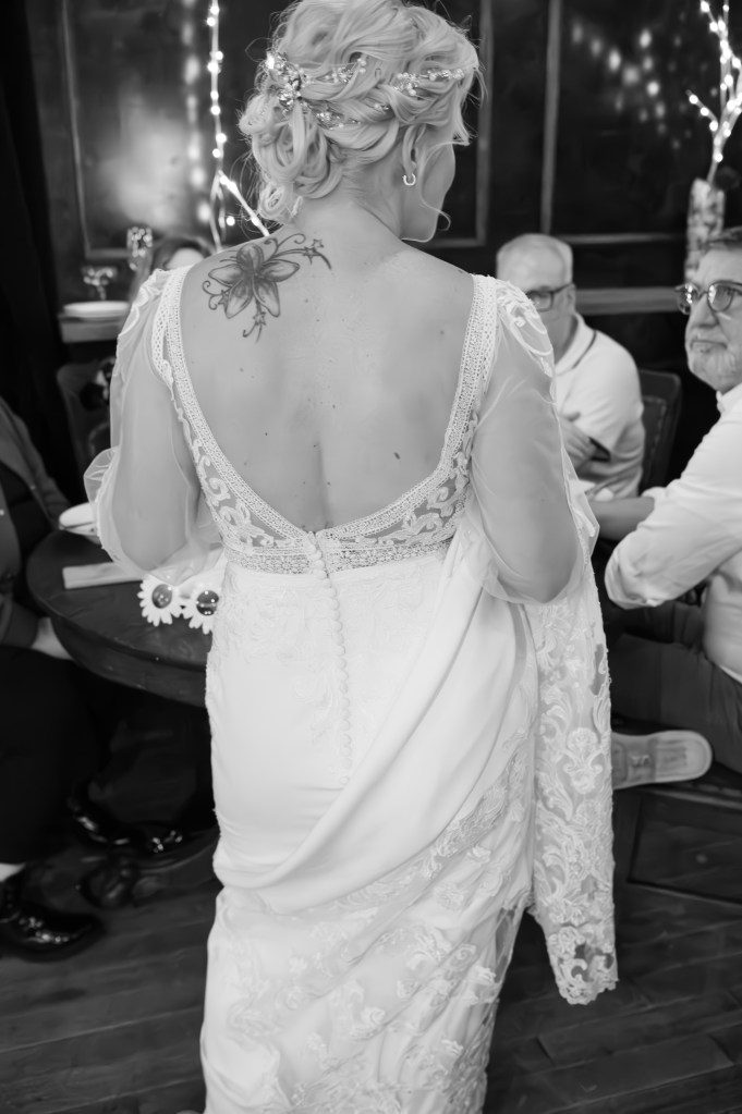 Une femme en robe de mariée tournée de dos, mettant en valeur son tatouage sur l'épaule et des détails élégants de sa tenue. En arrière-plan, des invités assis et une ambiance festive.