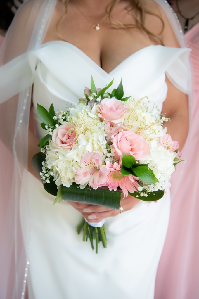Une mariée tenant un bouquet de fleurs, avec des roses roses et blanches, entourées de verdure, vêtue d'une robe blanche avec un voile.