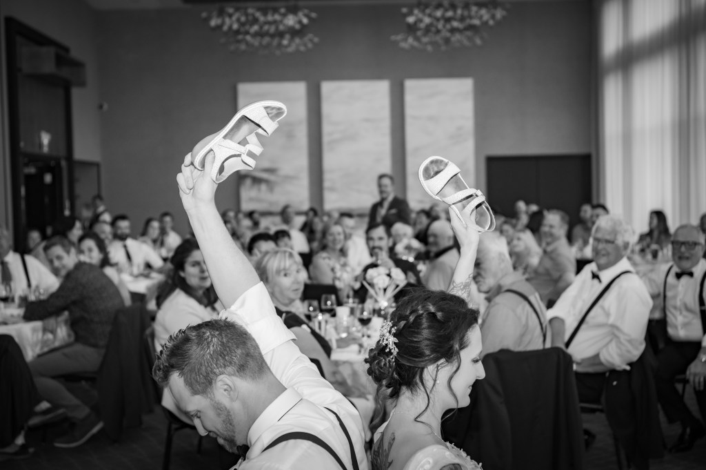 Un couple de mariés levant chacun une chaussure lors d'une réception de mariage, entourés de convives souriants dans la salle de réception à l'Espace 4 Saison à Orford.