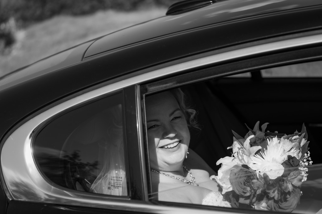 Une mariée souriante à l'intérieur d'une voiture, tenant un bouquet de fleurs, en noir et blanc.