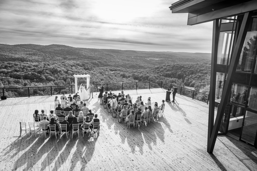 Cérémonie de mariage en plein air, avec un couple devant une arche, entouré d'invités assis sur une terrasse surplombant des collines verdoyantesà l'auberge de la Montagne Coupée.