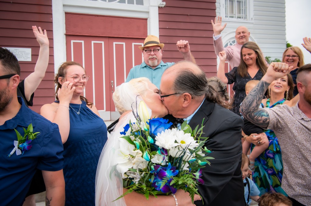 Un couple s'embrasse devant un bâtiment, entouré de leurs amis et famille célébrant leur mariage.