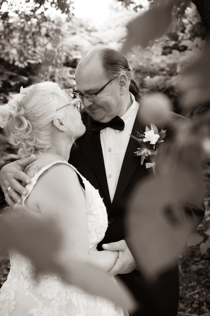Un couple âgé s'embrassant tendrement en noir et blanc, entouré de verdure.