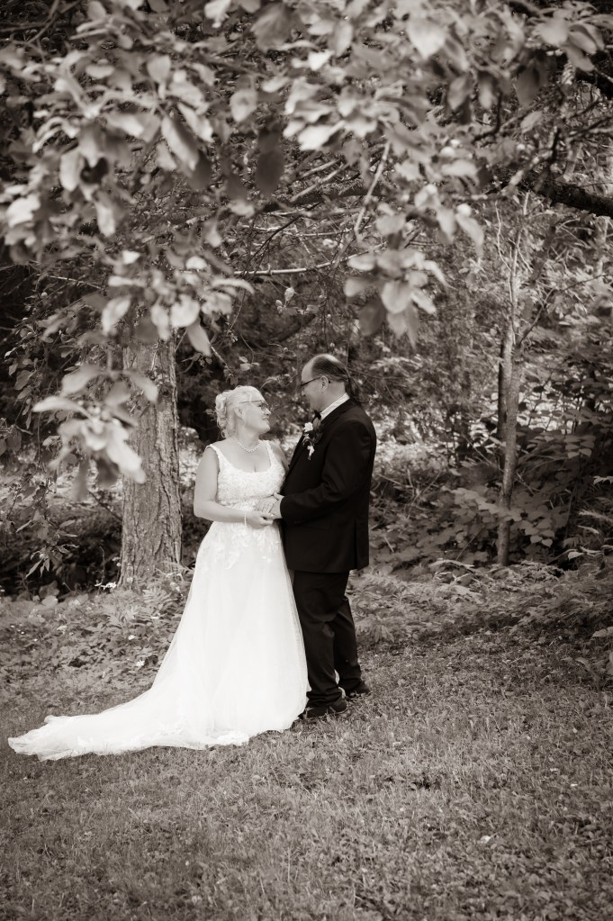 Un couple se regarde tendrement sous un arbre, le marié en costume noir et la mariée en robe blanche, dans un cadre naturel.