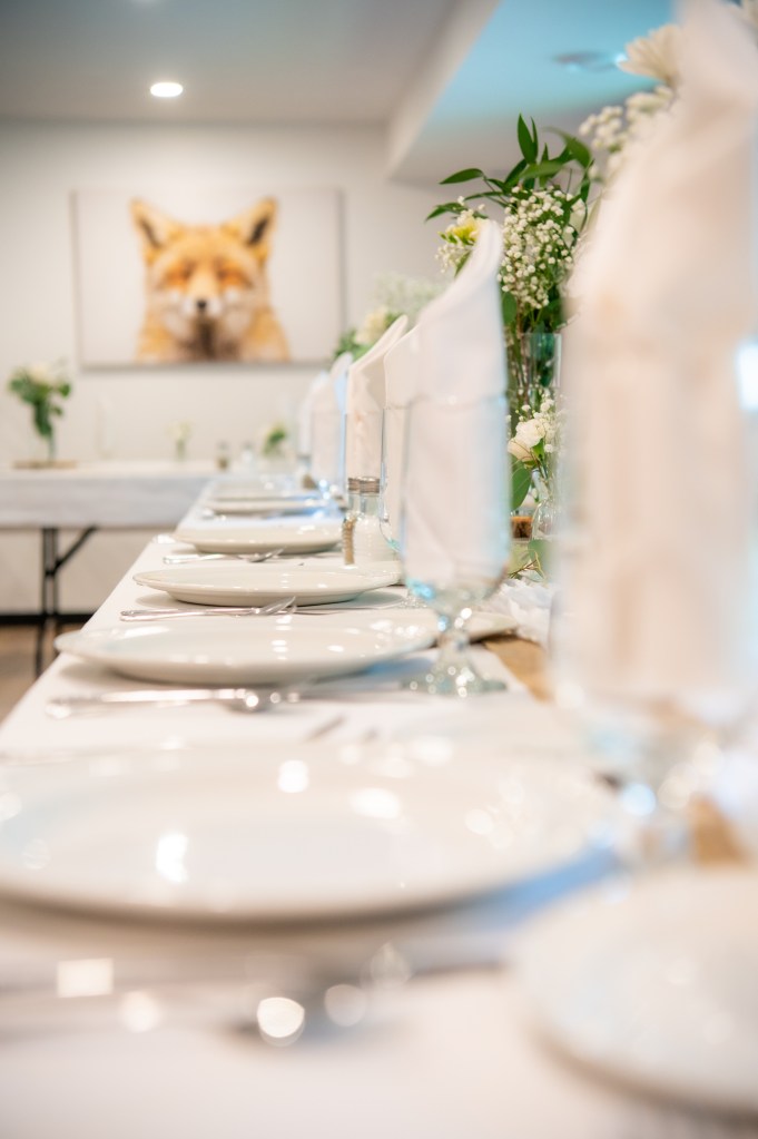 Table dressée avec des assiettes blanches, verres et serviettes en papier, décorée de fleurs et d'une œuvre d'art d'un renard en arrière-plan.