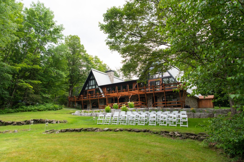 Maison de style chalet entourée d'arbres, avec une grande véranda et des chaises blanches disposées sur la pelouse.