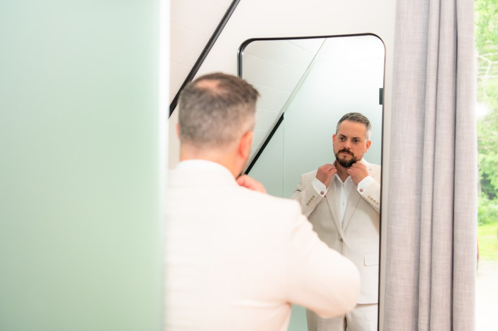 Un homme en costume clair ajuste sa cravate devant un miroir.