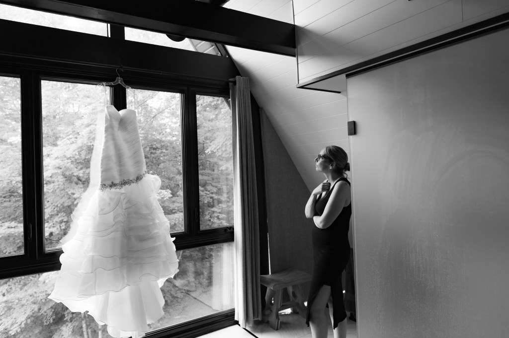 Une mariée contemplant sa robe de mariée accrochée près d'une fenêtre, avec un paysage naturel en arrière-plan.