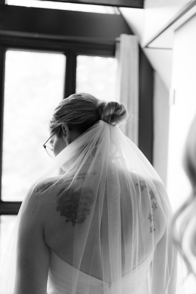 Femme en robe de mariée vue de dos, portant un voile et des lunettes, avec des tatouages visibles sur les épaules.