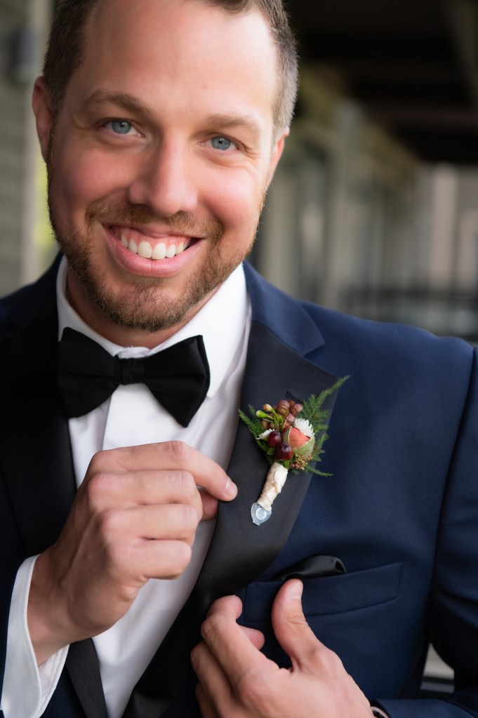 Un homme souriant en costume sombre avec un nœud papillon, ajustant sa boutonnière décorée de fleurs et de feuillages.