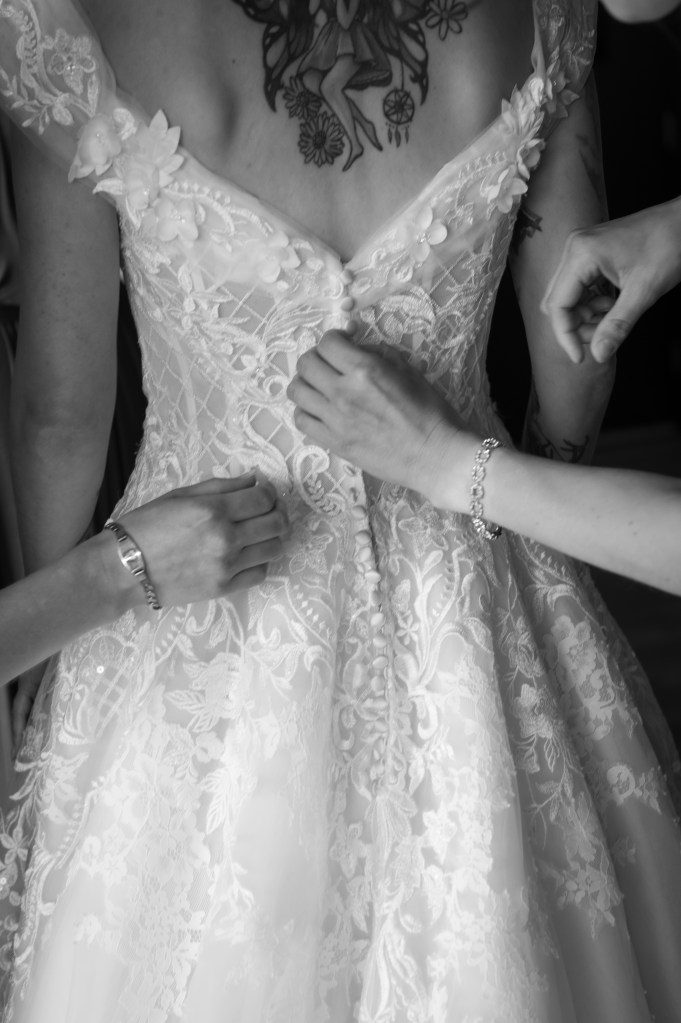 Une mariée en train de se préparer, avec des mains aidant à fermer le dos de sa robe de mariée ornée de dentelle.
